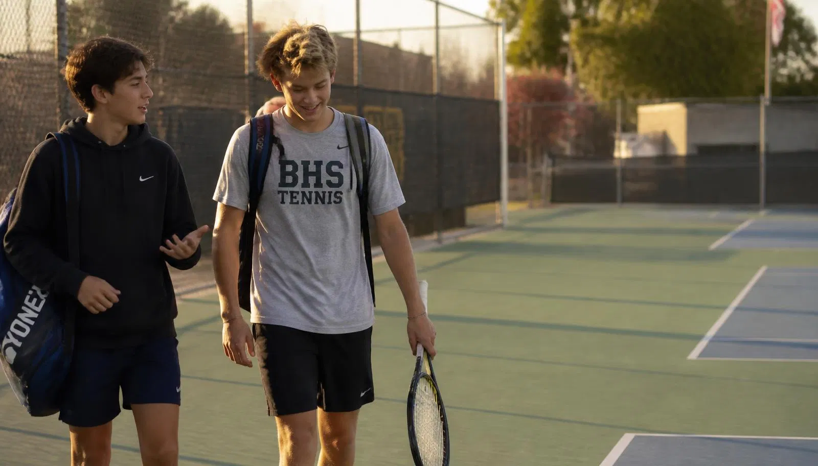 A high school tennis player walking off an outdoor school court with a friend after practice in late-afternoon light