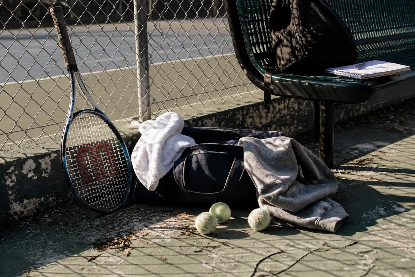 A tennis racquet, bag and towel left by the fence on a worn outdoor school court
